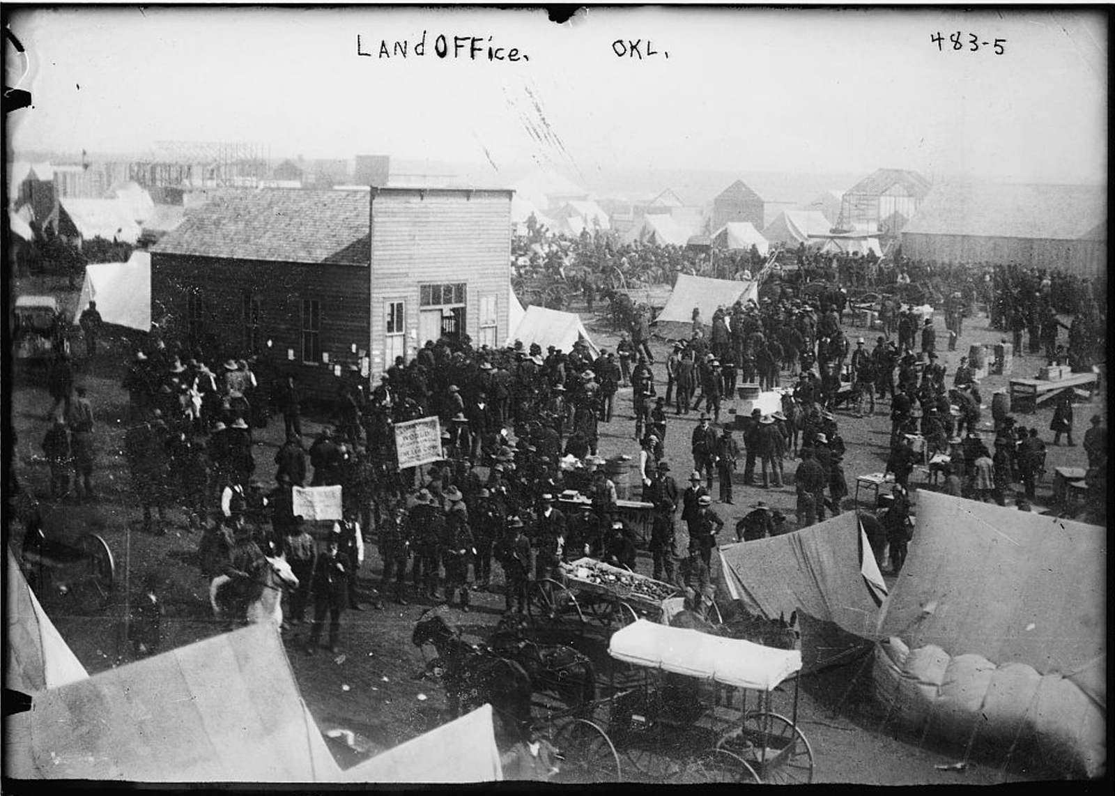 A crowd of people, wagons and tents gathered around a wooden land office building.